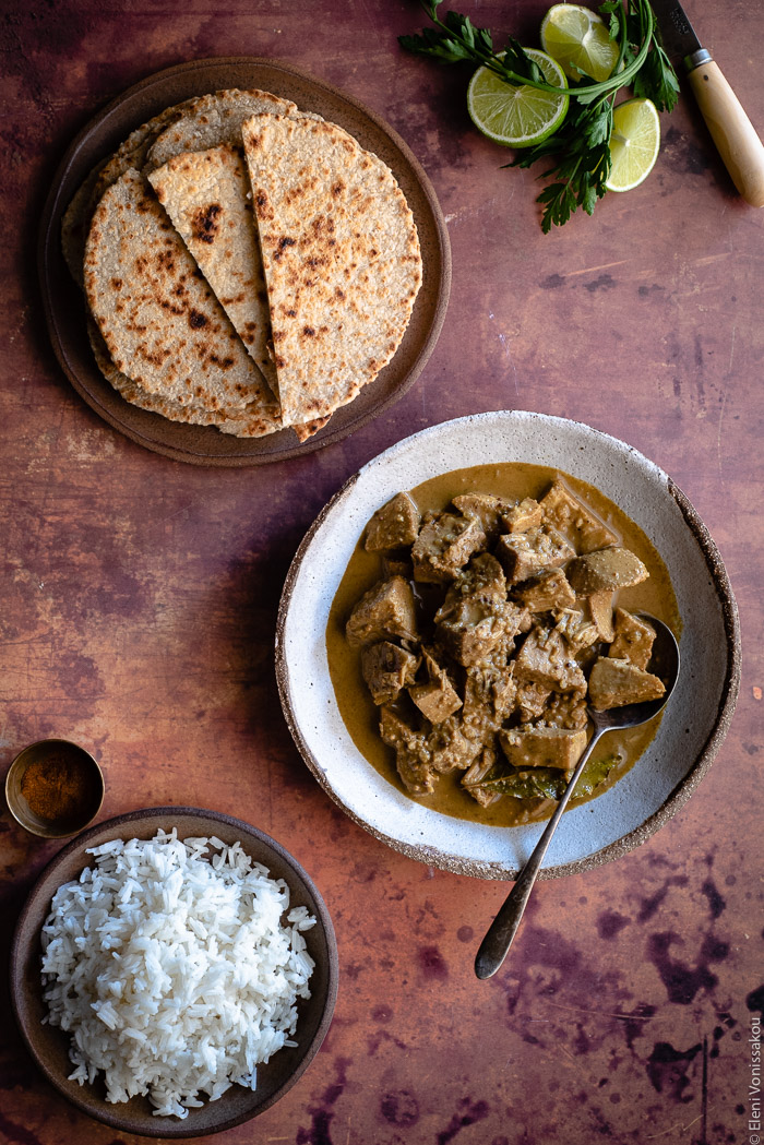 Sri Lankan Inspired Jackfruit Curry in the Slow Cooker www.thefoodiecorner.gr Photo description: A bowl of jackfruit curry in sauce in the middle of the image. To the bottom a plate of fluffy cooked basmati rice and to the top a plate of stacked flatbreads. Barely visible in the top right corner some cut lime and parsley.