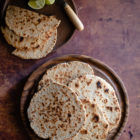 Sri Lankan Inspired Coconut Flatbread (Pol Roti) with Desiccated Coconut www.thefoodiecorner.gr Photo description: A pile of round coconut flatbreads (roti) on a wooden chopping board. To the top left is a small plate with a few cut pieces of flatbread, some lime wedges and a knife.