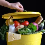 12 Ways To Easily Reduce Food Waste At Home www.thefoodiecorner.gr Photo description: A yellow metal bin overflowing with vegetables, fruit, milk and cheese. A hand is holding the lid just above the food in the bin.