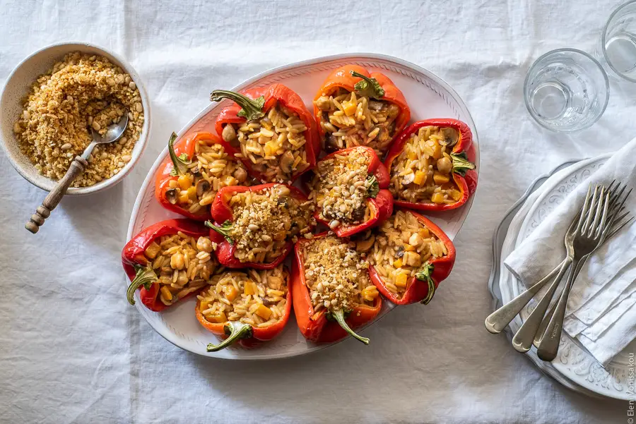 Roasted Red Peppers Stuffed with Mushroom, Pumpkin and Harissa Chickpea Orzotto www.thefoodiecorner.gr Photo description: A platter full of roasted red pepper halves stuffed with orzotto, on a white linen tablecloth. To the left of the image a small bowl with breadcrumb and pine nut topping. To the right, half visible, a stack of plates with some cutlery and two small glasses.