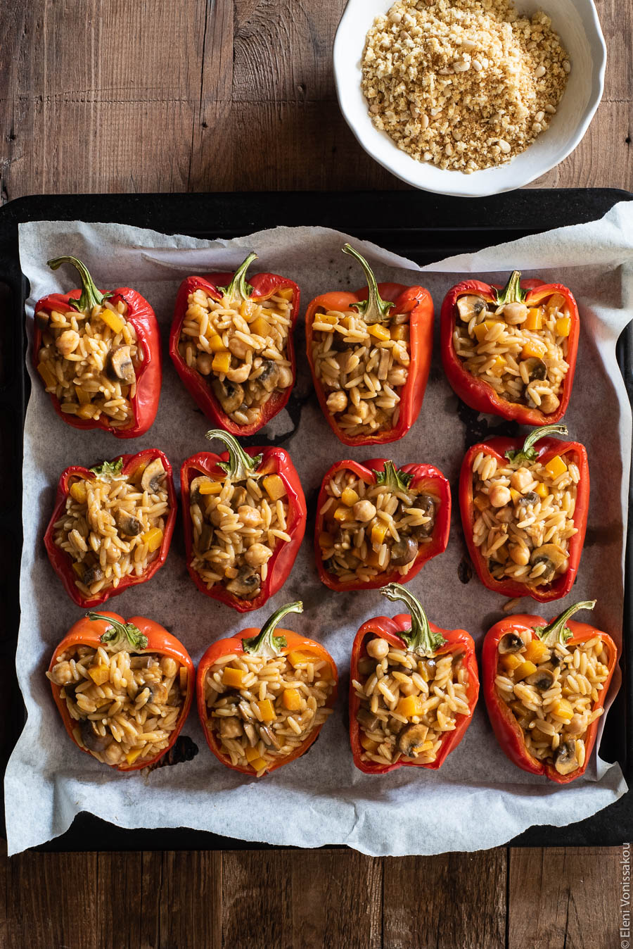 Roasted Red Peppers Stuffed with Mushroom, Pumpkin and Harissa Chickpea Orzotto www.thefoodiecorner.gr Photo description: A baking tray lined with grease proof paper and filled with red pepper halves stuffed with orzotto. Above the tray is a bowl with breadcrumb and pine nut topping.