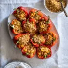 Roasted Red Peppers Stuffed with Mushroom, Pumpkin and Harissa Chickpea Orzotto www.thefoodiecorner.gr Photo description: A platter full of roasted red pepper halves stuffed with orzotto, on a white linen tablecloth. To the top of the image a small bowl with breadcrumb and pine nut topping. To the bottom, half visible, a stack of plates with some cutlery and two small glasses.