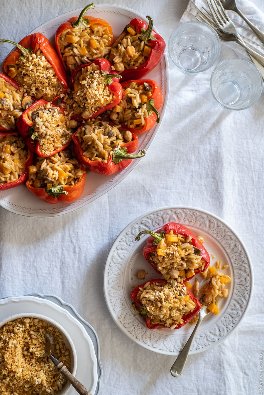 Roasted Red Peppers Stuffed with Mushroom, Pumpkin and Harissa Chickpea Orzotto www.thefoodiecorner.gr Photo description: Laid out on a linen tablecloth are a platter with orzotto-stuffed red peppers, two small glasses, a plate with a couple of stuffed peppers and a fork on it, and (barely visible) a small bowl with breadcrumb and pine nut topping. 